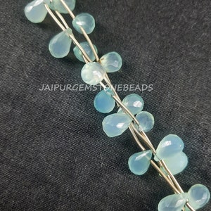 May include: A strand of light blue gemstone beads in a teardrop shape. The beads are strung on silver wire and are arranged in a cascading pattern. The beads are on a dark gray background.  JAIPURGEMSTONEBEADS