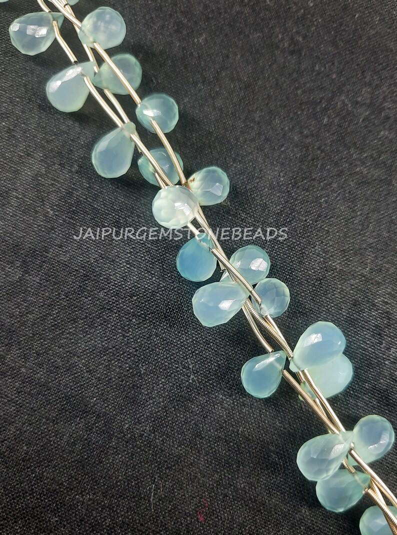 May include: A strand of light blue gemstone beads in a teardrop shape. The beads are strung on silver wire and are arranged in a cascading pattern. The beads are on a dark gray background.  JAIPURGEMSTONEBEADS
