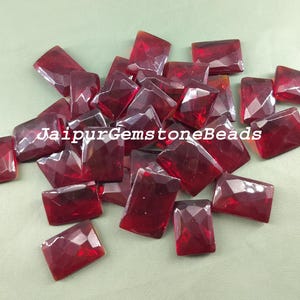 Puede incluir: Un montón de gemas de vidrio rectangulares rojas facetadas. Las gemas están esparcidas sobre una superficie verde. JaipurGemstoneBeads está escrito en texto blanco en la imagen.