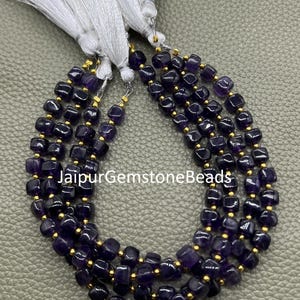 May include: Dark purple gemstone beads with gold spacers, strung together in multiple strands. The beads are square-shaped. White tassels are visible at the top. The text "JaipurGemstoneBeads" is printed on the image.