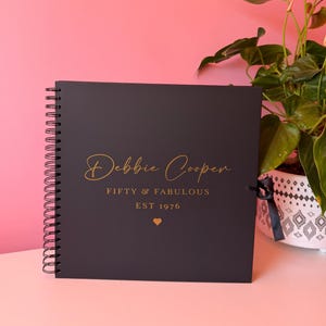 Könnte beinhalten: Ein quadratisches, schwarzes Notizbuch mit Spiralbindung und goldfarbenem Text, der Debbie Cooper, FIFTY & FABULOUS, EST 1976 sowie ein kleines goldenes Herz zeigt. Das Notizbuch hat ein schwarzes Schleifenband an der rechten Seite.
