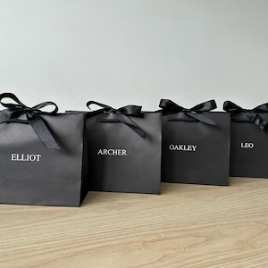 May include: Four black gift bags tied with black satin bows. Each bag features a name in white: Elliot, Archer, Oakley, and Leo. The bags are displayed on a light wooden surface.