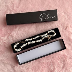 May include: A necklace made of small white shell beads and gold-toned metal beads, featuring a gold-toned starfish charm. The necklace is displayed inside a black rectangular box with the name Olivia printed in white script on the lid.