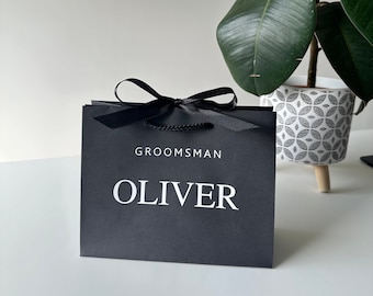 May include: A black gift bag with a black satin ribbon and bow. The bag has the words "Groomsman Oliver" printed in white on the front.