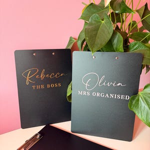 Könnte beinhalten: Drei schwarze Klemmbretter mit Metallklemmen. Eines trägt den goldenen Schriftzug Rebecca THE BOSS, ein anderes den weißen Schriftzug Olivia MRS ORGANISED und das dritte ist leer. Die Klemmbretter stehen vor einer rosa Wand.