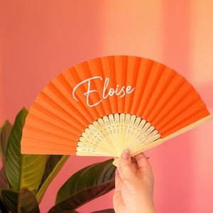 May include: A bright orange hand fan featuring a light-colored wooden frame with intricate cut-out details. The name Eloise is printed in a white cursive script across the center of the pleated fabric surface of the folding hand fan.