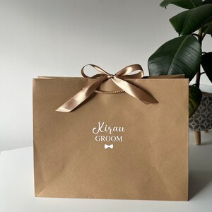 Pode incluir: Uma sacola de presente de papel castanho com uma fita e um la&ccedil;o dourados. A sacola tem o texto "Kiran GROOM" impresso em branco na frente com um pequeno &iacute;cone de gravata borboleta.