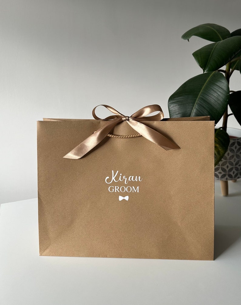 Pode incluir: Uma sacola de presente de papel castanho com uma fita e um la&ccedil;o dourados. A sacola tem o texto "Kiran GROOM" impresso em branco na frente com um pequeno &iacute;cone de gravata borboleta.