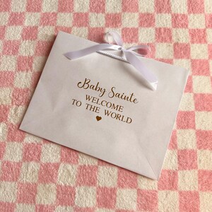 May include: A white gift bag with a white ribbon bow, featuring the text "Baby Sainte WELCOME TO THE WORLD" in gold script. A small gold heart is below the text. The bag rests on a pink and white checkered surface.