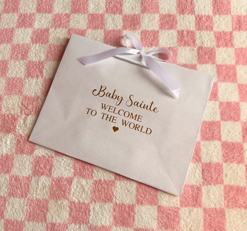 May include: A white gift bag with a white ribbon bow, featuring the text "Baby Sainte WELCOME TO THE WORLD" in gold script. A small gold heart is below the text. The bag rests on a pink and white checkered surface.