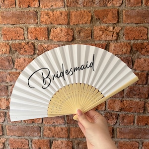 May include: A white folding hand fan with a light beige wooden frame featuring intricate cut-out details. The word "Bridesmaid" is printed in a black cursive script across the center of the white fabric surface of the open fan.