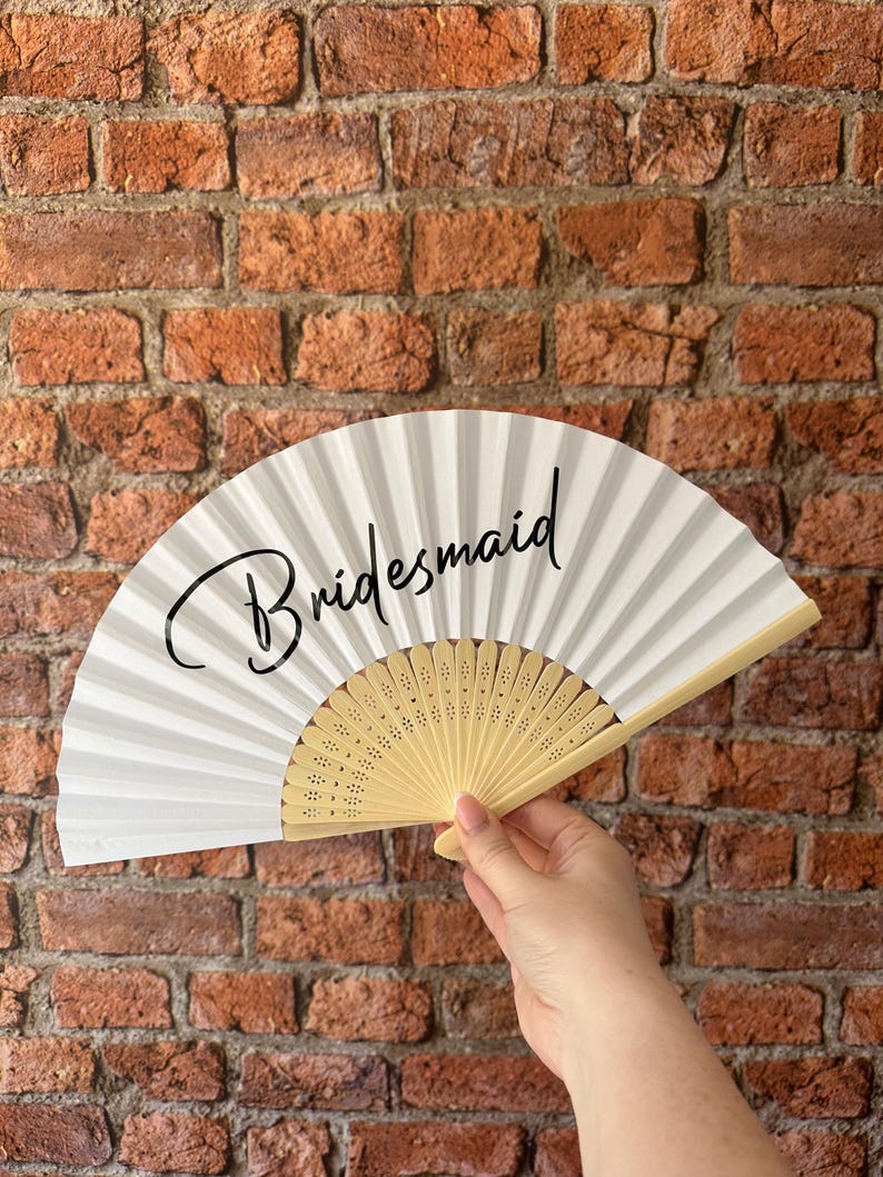 May include: A white folding hand fan with a light beige wooden frame featuring intricate cut-out details. The word "Bridesmaid" is printed in a black cursive script across the center of the white fabric surface of the open fan.