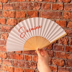 May include: A white folding hand fan with a light wood frame and the name Jasmine written in a red cursive script across the center of the fan surface. The fan is held open to display the pleated material and the wooden ribs at the base.