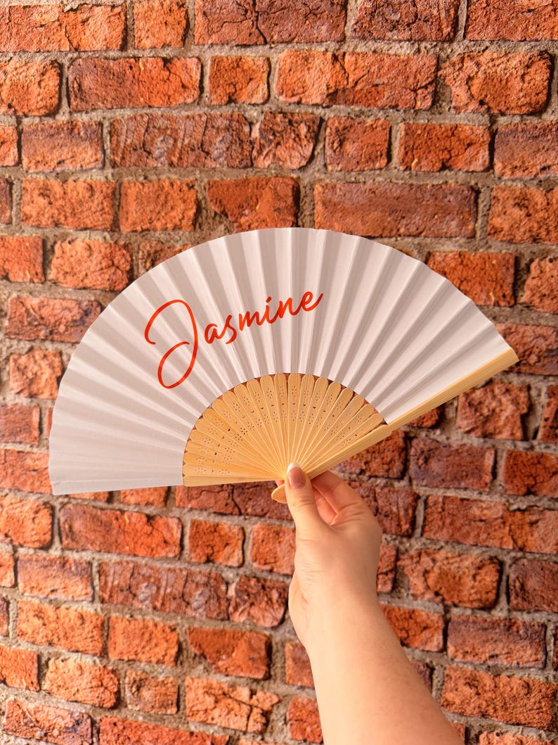 May include: A white folding hand fan with a light wood frame and the name Jasmine written in a red cursive script across the center of the fan surface. The fan is held open to display the pleated material and the wooden ribs at the base.