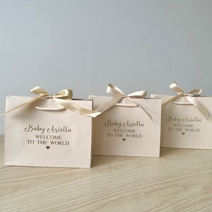 May include: Three beige gift bags with gold satin ribbon bows. Each bag has the text "Baby Ariella WELCOME TO THE WORLD" printed in a dark brown script font, with a small heart symbol below.