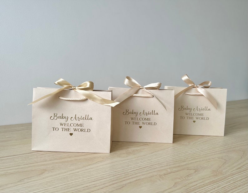 May include: Three beige gift bags with gold satin ribbon bows. Each bag has the text "Baby Ariella WELCOME TO THE WORLD" printed in a dark brown script font, with a small heart symbol below.