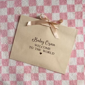 May include: A beige gift bag with a peach-colored ribbon bow. The bag has the text "Baby Oran WELCOME TO THE WORLD" in black script. The background is a pink and white checkered pattern.