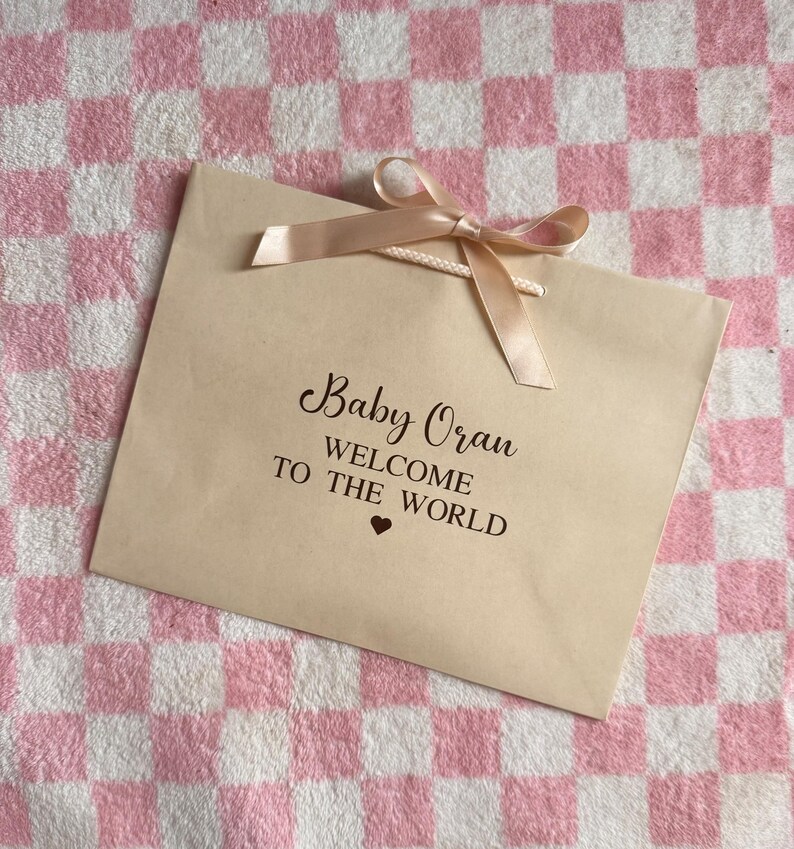 May include: A beige gift bag with a peach-colored ribbon bow. The bag has the text "Baby Oran WELCOME TO THE WORLD" in black script. The background is a pink and white checkered pattern.