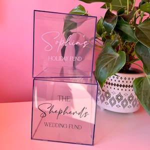 May include: Two stacked clear acrylic square boxes with slots on top. The top box features white text reading Sophia's HOLIDAY FUND. The bottom box features black text reading THE Shepherd's WEDDING FUND. Both boxes are transparent with visible seams.