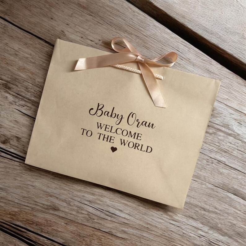 May include: A tan gift bag with a peach-colored ribbon tied in a bow. The bag has the words "Baby Oran WELCOME TO THE WORLD" printed in black script, with a small heart symbol below.