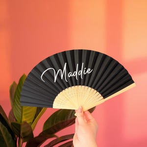 May include: A black folding hand fan with a light-colored wooden frame and the name Maddie written in white cursive script across the center of the pleated fabric. The fan is fully extended to display its semi-circular shape and design.