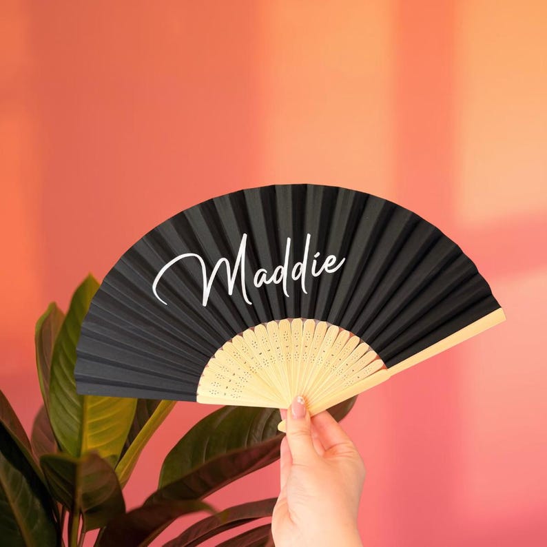 May include: A black folding hand fan with a light-colored wooden frame and the name Maddie written in white cursive script across the center of the pleated fabric. The fan is fully extended to display its semi-circular shape and design.