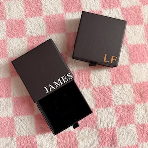 May include: Two dark brown gift boxes with black interiors are displayed on a pink and white checkered surface. One box is open, revealing a black interior, with the name "JAMES" printed in white. The other box is closed, with the initials "LF" in gold.