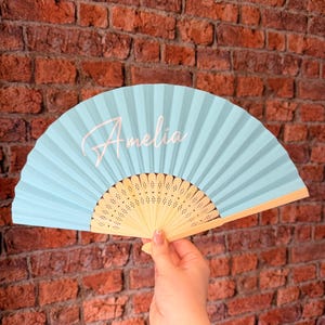 May include: A light blue handheld folding fan with a wooden frame featuring intricate cut-out patterns. The name Amelia is printed in a white cursive script on the left side of the pleated fabric surface of the fan.