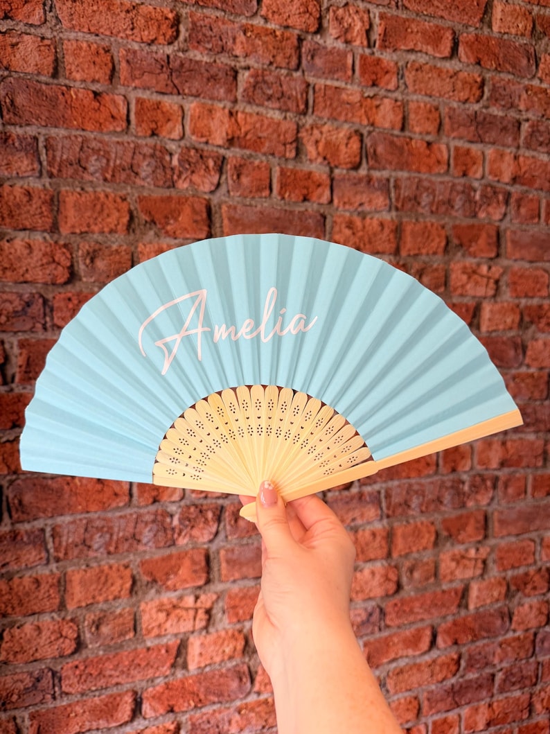 May include: A light blue handheld folding fan with a wooden frame featuring intricate cut-out patterns. The name Amelia is printed in a white cursive script on the left side of the pleated fabric surface of the fan.