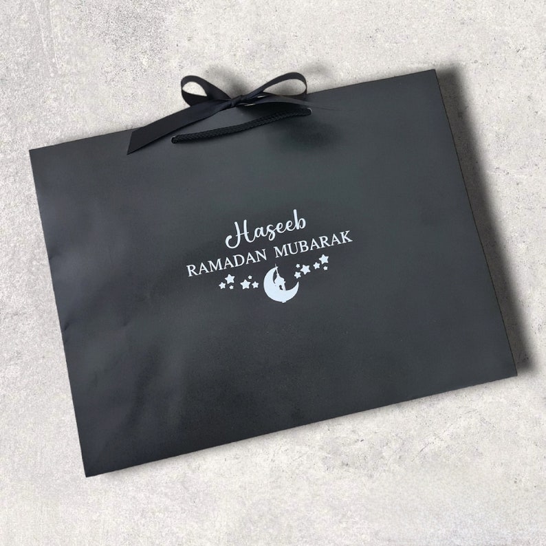 May include: Black gift bag with a black ribbon and a white design that says "Haseeb Ramadan Mubarak" with a crescent moon and stars.