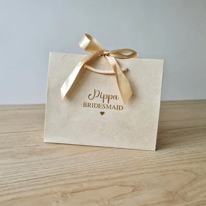 Pode incluir: Uma sacola de presente marrom claro com uma fita dourada e o texto "Dippa Bridesmaid" em letras douradas.