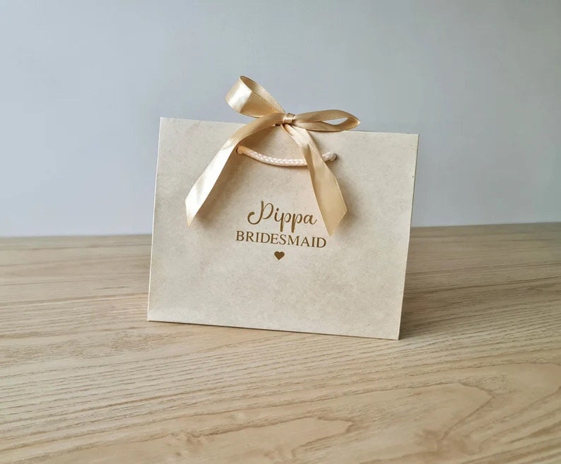 Pode incluir: Uma sacola de presente marrom claro com uma fita dourada e o texto "Dippa Bridesmaid" em letras douradas.