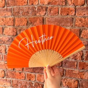 May include: A bright orange hand fan featuring the name Heather written in white cursive script across the center. The fan is constructed with a pleated fabric canopy and a light-colored wooden frame with decorative cutouts at the base.