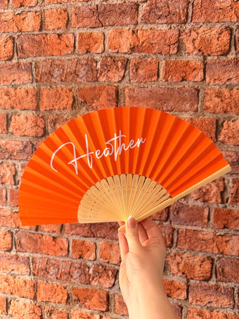 May include: A bright orange hand fan featuring the name Heather written in white cursive script across the center. The fan is constructed with a pleated fabric canopy and a light-colored wooden frame with decorative cutouts at the base.