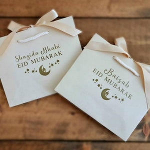 May include: Two white gift bags with gold ribbon and gold lettering. The bags are decorated with a crescent moon and stars. The text on the bags reads "Shazida Bhabi Eid Mubarak" and "Baisaib Eid Mubarak".