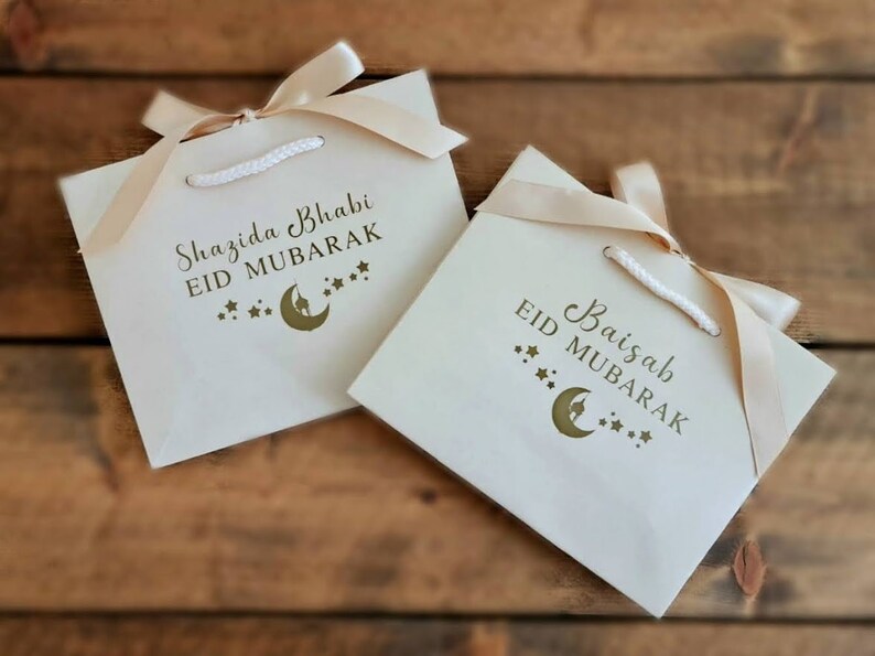May include: Two white gift bags with gold ribbon and gold lettering. The bags are decorated with a crescent moon and stars. The text on the bags reads "Shazida Bhabi Eid Mubarak" and "Baisaib Eid Mubarak".