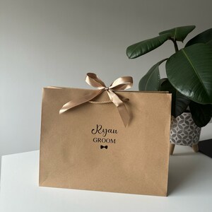 Pode incluir: Uma sacola de presente de papel castanho com uma fita e um la&ccedil;o dourados. A sacola &eacute; personalizada com o nome "Ryan" e a palavra "Groom" com um s&iacute;mbolo de gravata borboleta.