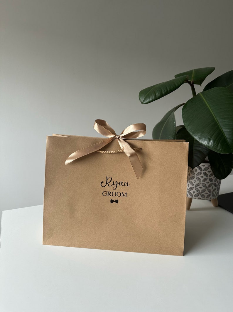 Pode incluir: Uma sacola de presente de papel castanho com uma fita e um la&ccedil;o dourados. A sacola &eacute; personalizada com o nome "Ryan" e a palavra "Groom" com um s&iacute;mbolo de gravata borboleta.