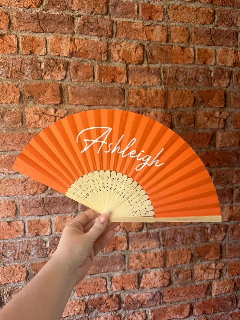 May include: A bright orange folding hand fan featuring the name Ashleigh written in white cursive script across the center. The fan has a light-colored wooden frame with decorative cut-out patterns near the base of the ribs.