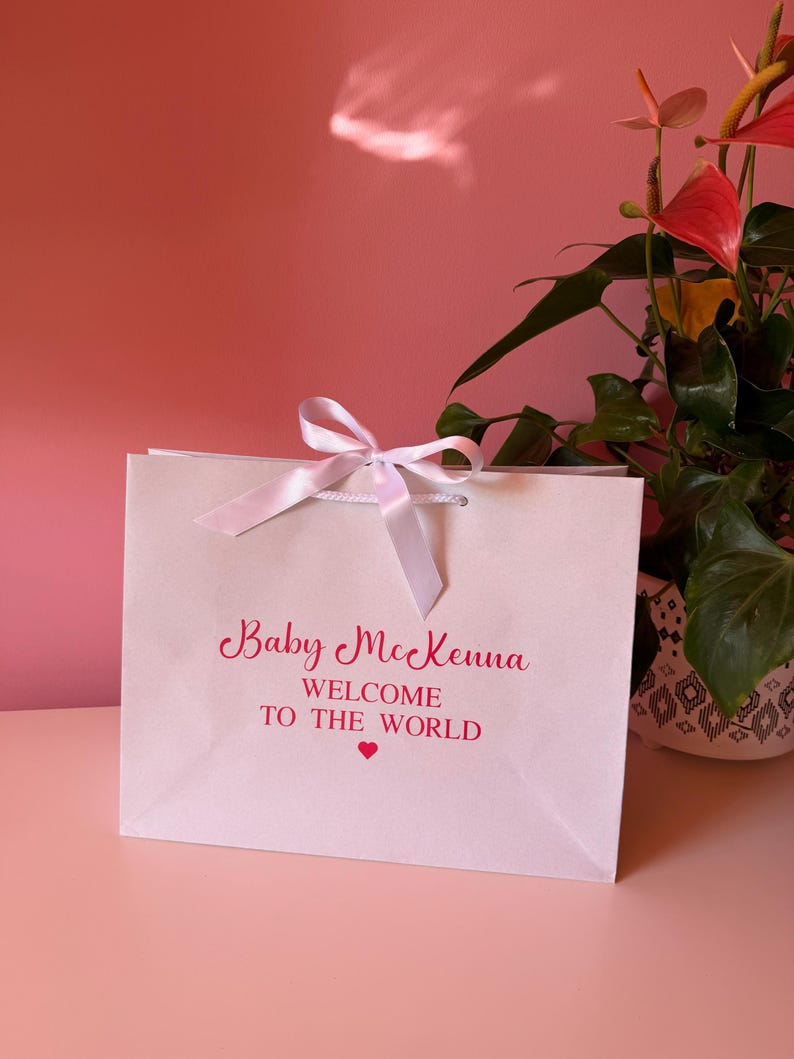 May include: A white gift bag with a pink ribbon tied in a bow. The bag has the text "Baby McKenna WELCOME TO THE WORLD" in pink script. A small pink heart is below the text. The bag is on a pink surface.