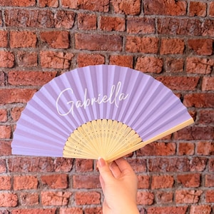 May include: A handheld folding fan featuring a light purple fabric canopy with the name Gabriella printed in white script lettering. The fan has a light-colored wooden frame with intricate laser-cut decorative patterns along the base ribs.