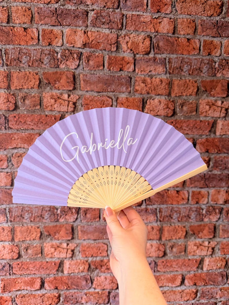 May include: A handheld folding fan featuring a light purple fabric canopy with the name Gabriella printed in white script lettering. The fan has a light-colored wooden frame with intricate laser-cut decorative patterns along the base ribs.