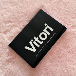 Könnte beinhalten: Ein schwarzes Notizbuch mit dem Wort "Vitori" in großen weißen Buchstaben und dem Text "ADVISORY | OPERATOR" darunter. Das Notizbuch liegt auf einer rosa, flauschigen Oberfläche.