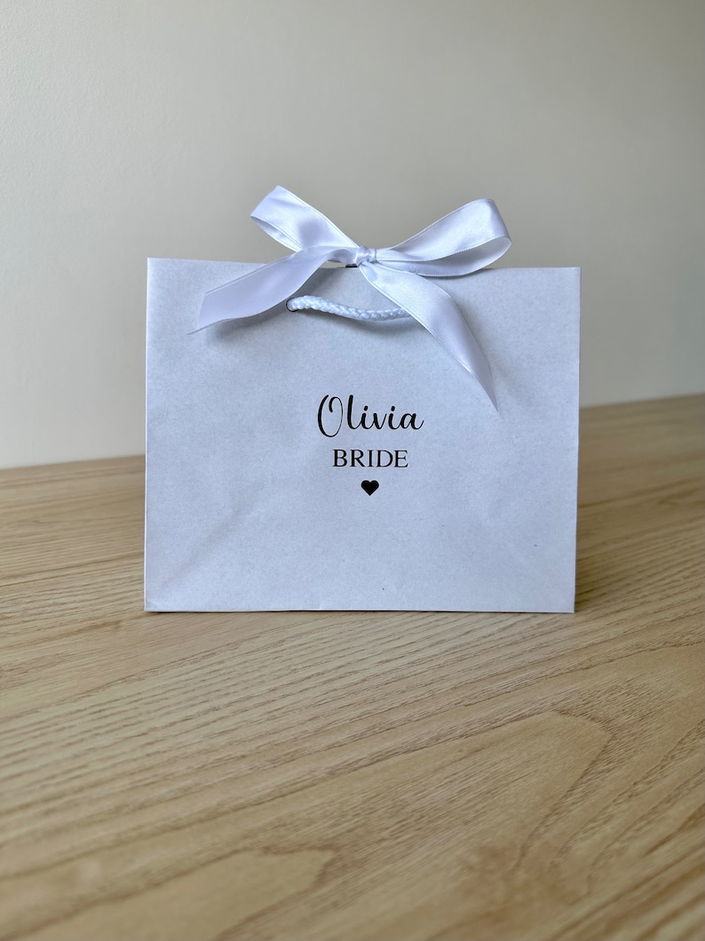 Pode incluir: Uma sacola de presente branca com um la&ccedil;o de fita de cetim branco. A sacola tem o texto "Olivia Bride" impresso na frente com um pequeno cora&ccedil;&atilde;o preto.