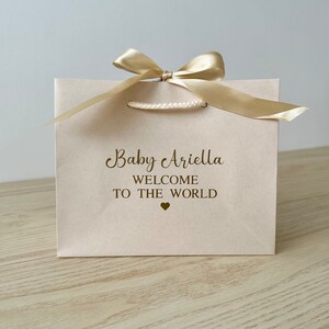 May include: A light pink gift bag with a gold satin bow and handles. The bag has the text "Baby Ariella WELCOME TO THE WORLD" printed in gold. A small gold heart is below the text. The bag is on a light wood surface.