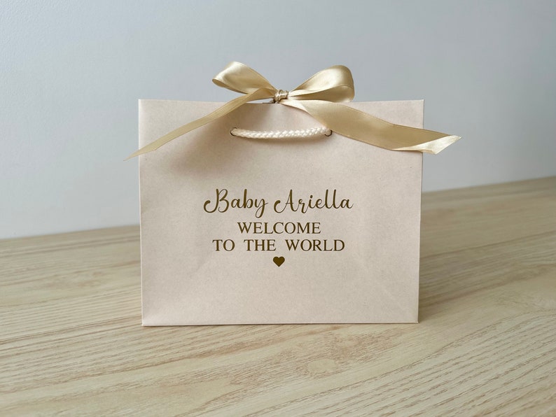 May include: A light pink gift bag with a gold satin bow and handles. The bag has the text "Baby Ariella WELCOME TO THE WORLD" printed in gold. A small gold heart is below the text. The bag is on a light wood surface.