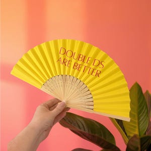 May include: A bright yellow folding hand fan with a light-coloured wooden frame. The fan features the text "DOUBLE DS ARE BETTER" printed in a dark red font across the pleated surface. The fan is held open to display the full semi-circular shape.