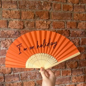 May include: A bright orange hand fan featuring the black text I'm on holiday printed across the center. The fan has a pleated fabric surface and a light-colored, perforated plastic frame that radiates from the base in a semi-circular shape.