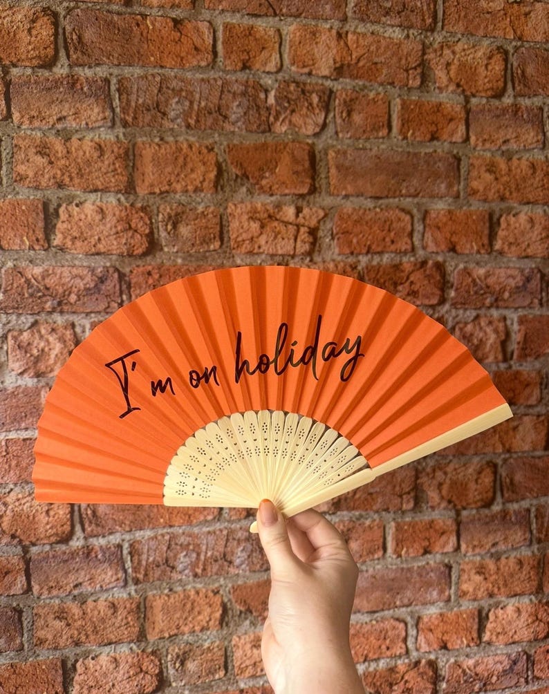 May include: A bright orange hand fan featuring the black text I'm on holiday printed across the center. The fan has a pleated fabric surface and a light-colored, perforated plastic frame that radiates from the base in a semi-circular shape.