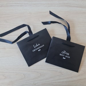 Pode incluir: Duas sacolas de presente pretas com texto branco e um s&iacute;mbolo de gravata borboleta. A sacola &agrave; esquerda diz "Luka Groomsman" e a sacola &agrave; direita diz "Ross Best Man".